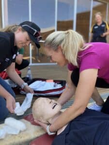 Two blonde women, one with a baseball hat and sunglasses, and another in a fuschia shirt, practice stabilising a lifelike CPR mannequin.